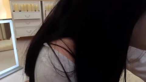 Media: Video of a young woman with long, straight black hair, partially obscuring her face, wearing a white shirt. Background features a beige carpet, white dresser with drawers, and a mirror.