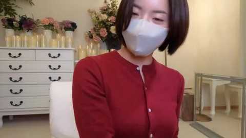 Media: Video of an Asian woman with short dark hair, wearing a white face mask, a red cardigan, and a white chair, in a room with a white dresser, glass table, and floral arrangement.