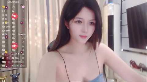 Media: Video of a young East Asian woman with long black hair, fair skin, and a slender physique, wearing a light blue spaghetti-strap top, standing indoors with beige curtains and a flat-screen TV in the background.