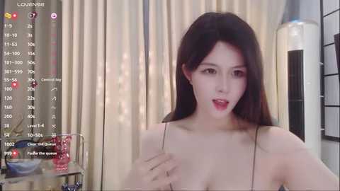 Media: A video of an East Asian woman with long, straight black hair, fair skin, and red lipstick, wearing a thin, black spaghetti strap top, posing in a bedroom with beige curtains and a tall, cylindrical lamp.