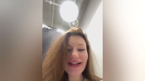 Media: A video captures a woman with long, straight brown hair, smiling with closed eyes, standing in a bright, modern room with a round light fixture and visible ceiling ductwork.