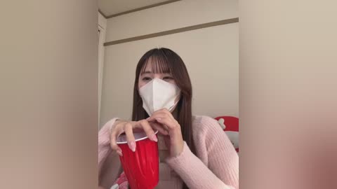 Media: Video of an East Asian woman with long brown hair and a white mask, holding a red cup, in a minimalist beige room with red and white pillows.