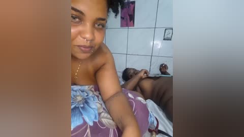 Media: A candid video of a topless Black woman with dark skin, large breasts, and a nose ring, lying next to a Black man with short hair on a bed with a floral blanket.