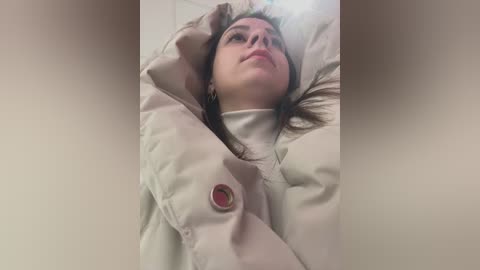 Media: Video of a young woman with light skin, brown hair, and a neutral expression, lying on her back, arms behind her head, wearing a white jacket with a red button.