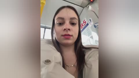 Media: A video of a young woman with long brown hair, wearing a beige coat, seated on a bus. She has a neutral expression. The background features bus interior details.