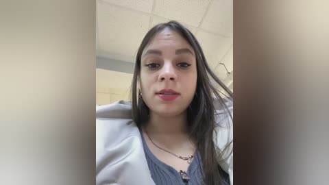 Media: Video of a young Latina woman with long dark hair, wearing a gray top, standing indoors in a dimly lit room with white ceiling tiles. She has a neutral expression.