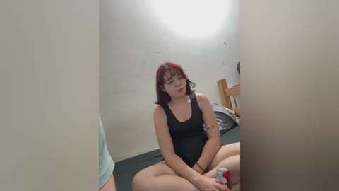 Media: Video of a young, fair-skinned woman with shoulder-length red hair and glasses, sitting cross-legged on a green floor in a small, dimly lit room with a white ceiling. She wears a black tank top and holds a can.