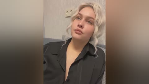 Media: A video of a young woman with fair skin and blonde hair, wearing a black satin robe, sitting in a room with a white wall and a power outlet.