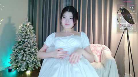 Media: A video of an East Asian woman with long dark hair in an off-the-shoulder white dress, sitting on a white couch in a dimly lit room with a lit Christmas tree, a clock, and gray curtains.