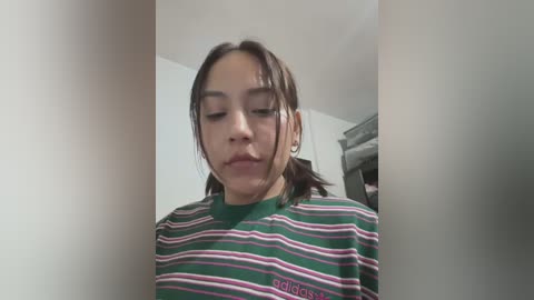 Media: Video of a young Asian woman with light skin, wearing a green and pink striped polo shirt, standing indoors. She has her eyes closed, looking contemplative.