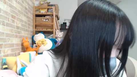 Media: A video of a young Asian girl with long black hair, wearing a white shirt, standing beside a brick wall filled with plush toys, including a bear and a fox.