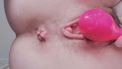 Video of a close-up of a person's shaved genital area with a pink, translucent dildo inserted into the vagina. The background is out of focus.