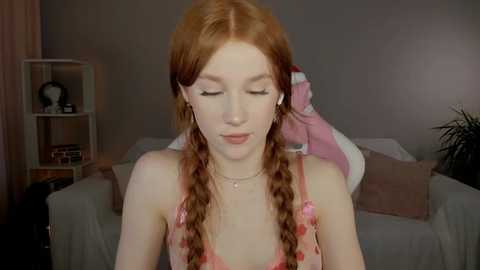 Media: Video of a fair-skinned, red-haired young woman with long braids, wearing a pink spaghetti strap top, seated in a dimly lit room with beige walls, a white couch, and a bookshelf.