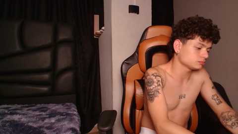 Media: Video of a shirtless young man with curly hair, tattoos, and white underwear, sitting on an orange gaming chair in a dimly lit bedroom with a black leather headboard and dark curtains.