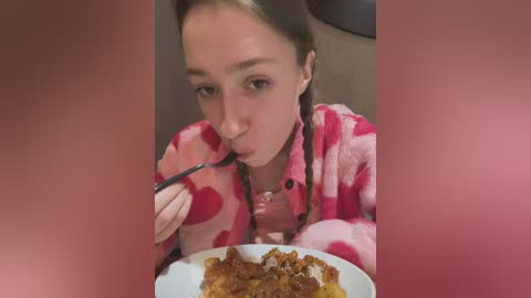 Media: A video shows a young Caucasian woman with fair skin, brown hair, and brown eyes, wearing a pink heart-patterned robe, eating from a white plate of spaghetti with a black fork. The background is blurred.