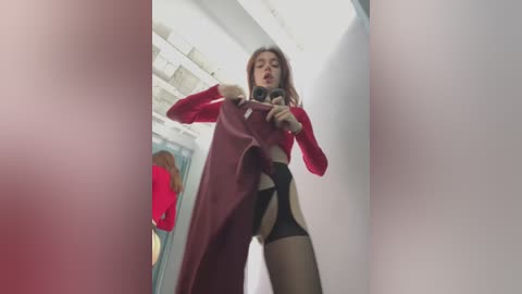 A video of a slender woman with fair skin and long brown hair, wearing a red long-sleeved top, black stockings, and a revealing red apron, taking a selfie in a brightly lit hallway.