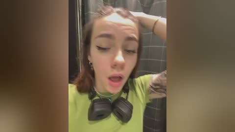 Media: Video of a young woman with light skin and brown hair, wearing a green shirt, black headphones, and a distressed expression, standing in a gray-tiled bathroom.