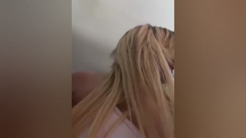 Media: A video of a blonde woman with long, straight hair, partially covering her face, wearing a light-colored top, standing in a dimly lit room with beige walls and a blurry background.