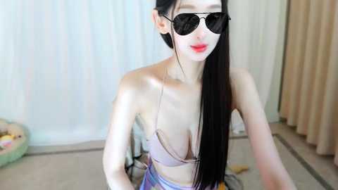 Media: Video of a slender, fair-skinned Asian woman with long black hair, wearing a strapless bra and reflective sunglasses, sitting on a bed in a room with white curtains and beige carpet.