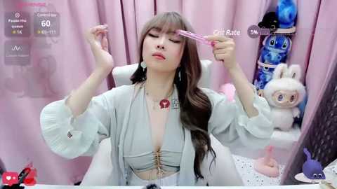 Media: Video of an Asian woman with long brown hair, wearing a light blue dress, applying eyeliner in a pink room with pastel decor, including stuffed animals.