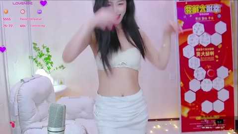 Media: Video of a slim Asian woman with long black hair, wearing a white bra and skirt, dancing energetically. Background includes a couch, a plant, and a festive red poster with Chinese text.