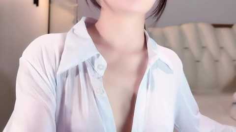 Video of an Asian woman with pale skin, dark hair tied back, wearing a sheer, light blue button-up shirt that is unbuttoned, revealing cleavage. Background features a beige, tufted headboard and a blurred, softly lit room.