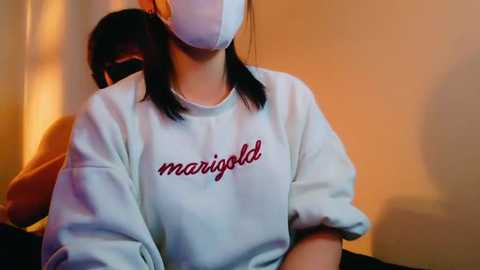 Media: Video of an East Asian woman in a white sweatshirt with \"marigold\" in red cursive, wearing a white face mask, seated indoors against a blurred, warm-toned background.