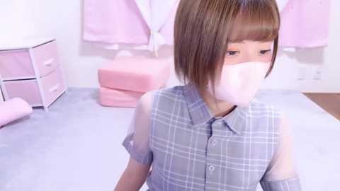 Media: Video of a young East Asian woman with short brown hair, wearing a plaid shirt, a white mask, and a light gray sweater, sitting in a pink room with pastel furniture and soft lighting.