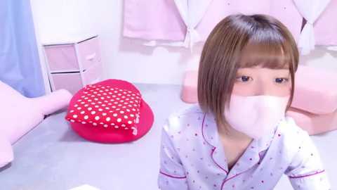 Media: Video of a young Asian girl with straight brown hair, wearing a white polka-dot pajama top and a white face mask, sitting on a light gray bed with pink heart pillow and pink quilt.