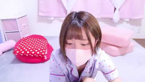 Media: A video of an East Asian child in a white mask, wearing a light blue pajama top, sitting on a pink bed with a red polka-dotted pillow, in a pastel-toned, organized bedroom.