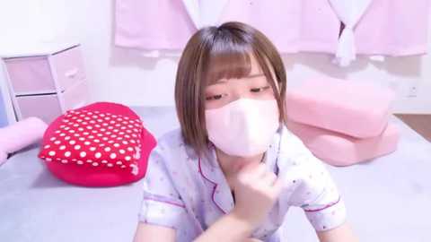 Media: Video of an Asian woman in a white, polka-dotted blouse, wearing a white face mask, sitting at a pink, heart-shaped cushioned desk, in a pastel-pink room with white walls.