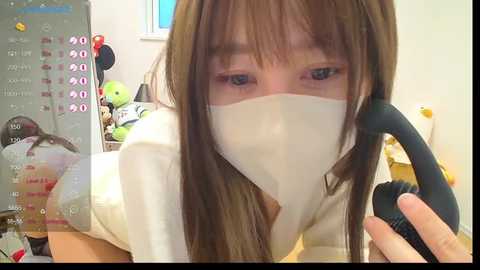 Media: Video of a young Asian woman with light brown hair, wearing a beige surgical mask and a white blouse, holding a black phone headset, in a cluttered room with stuffed toys and a bed.