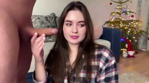 A video shows a young woman with long brown hair and pink lipstick kneeling, holding an erect penis, in a living room with a decorated Christmas tree and plaid shirt.