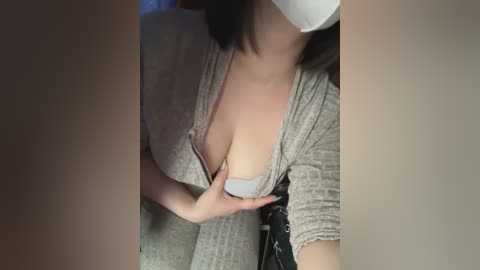 Media: Video of a woman with fair skin, wearing a grey knit top, revealing cleavage, and a white mask, taken from a slightly elevated angle.