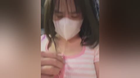 Video of a young girl with dark hair, wearing a pink striped shirt and white face mask, holding a smartphone, blurred background.