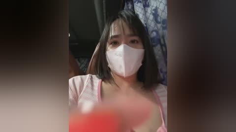 Media: A video of a woman with short black hair, wearing a white surgical mask and a pink top, sitting indoors with a blue patterned curtain in the background.