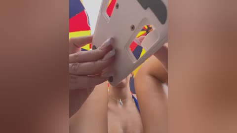 Video of a close-up, partially nude woman with light skin, holding a white phone with colorful flags, hands covered in red nail polish. Background includes a blue and yellow striped flag, suggesting a celebratory or political context.