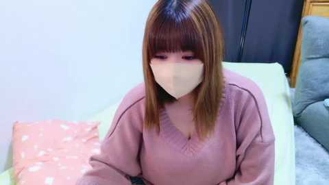 Media: Video of an Asian woman with straight, shoulder-length brown hair, wearing a beige face mask, pink sweater, and sitting on a bed with pink and gray pillows in a room with white walls and a black curtain.