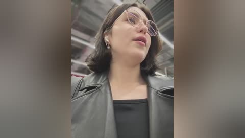 Media: A video of a young woman with light skin, wearing large glasses, a black jacket, and black top, taken indoors, with a blurred background.