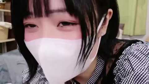 Video of a young Asian woman with straight black hair, wearing a white surgical mask, black and white striped shirt, and black lace top. She's indoors, with a blurred background showing a bed and green wall.