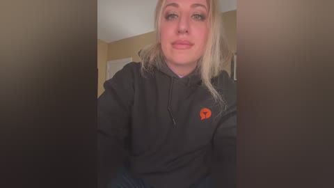 Media: Video of a blonde Caucasian woman with light skin, wearing a black hoodie with an orange logo and sitting indoors.