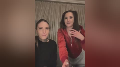 Media: Video of two young women with fair skin, smiling, one with long dark hair, red jacket, the other with black top, gray pants, both looking directly at the camera.