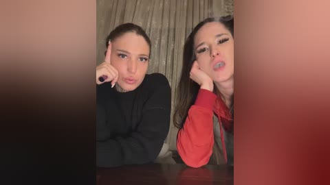 Media: Video of two women with fair skin, brown hair, and light makeup, leaning forward with pouty lips, wearing black and red sweaters, against a beige curtain backdrop.