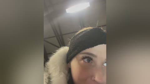 Media: Video of a woman with fair skin, wearing a black headband and a fur-trimmed coat, gazing to the right, in an industrial setting with a fluorescent light.