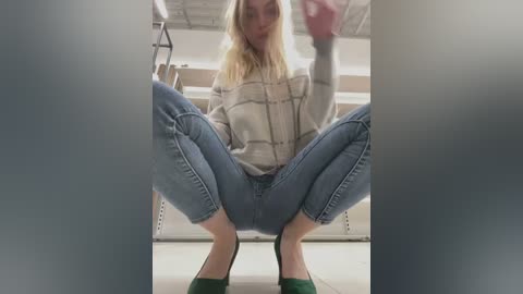 A video of a blonde woman squatting with her legs spread, wearing a beige jacket, blue jeans, and green shoes, indoors with a white ceiling and metallic shelving in the background.