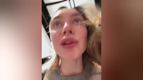 Media: Video of a Caucasian woman with light skin and blonde hair, wearing pink lipstick, looking up, slightly out of focus, against a blurred indoor background.