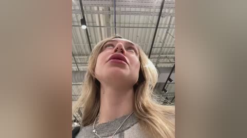 Video of a young woman with long blonde hair, fair skin, and closed eyes, looking upwards. She wears a gray sweater and a silver necklace. The background shows a modern, industrial ceiling with metal beams and lighting fixtures.