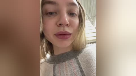 Video of a young woman with light skin, shoulder-length blonde hair, and a septum piercing, wearing a grey sweater, taken indoors with a blurry background.