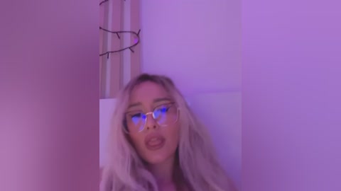 Media: Video of a young Caucasian woman with long blonde hair, wearing clear glasses and a light-colored sweater, standing against a white wall with purple lighting.