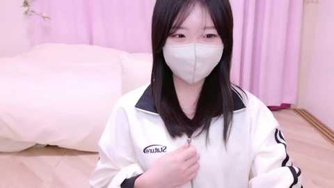 Media: A video of an East Asian woman with long black hair, wearing a white jacket and a mask, standing indoors with pastel-colored curtains and a bed in the background.